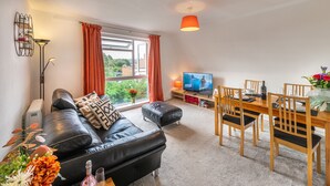 A living room with a black leather sofa, a wooden dining table with chairs, a TV, and a window with curtains. - Charming 2-bed Apartment in Windsor - Free Parking (Windsor)