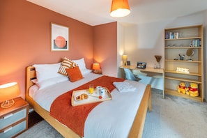 Een opgemaakt bed met een dienblad ontbijt, een bureau met een computer, en een boekenkast met boeken en decoratieve objecten.