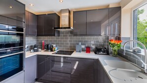 A modern kitchen with dark cabinetry, a built-in oven, a stainless steel range hood, and a large window providing natural light. - Charming 2-bed Apartment in Windsor - Free Parking (Windsor)