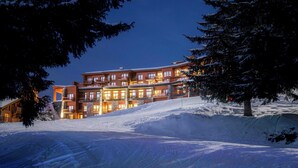 Ein mehrstöckiges Hotel inmitten einer verschneiten Landschaft, mit einem markanten Gebäude, mehreren Balkonen und einer Skipiste im Vordergrund.