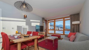 Ein modernes Wohnzimmer mit einem Holzespeisetisch, roten Stühlen, einem grauen Sofa und einem großen Fenster mit Blick auf die Berge.