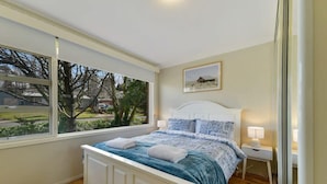 A bedroom with a large window, a bed with a blue bedspread, a white headboard, and a nightstand with a lamp. - Collwood Cottage. Four Beds + Fireplace. Orange. (Orange)