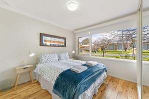 A bedroom with a bed, a bedside table, a lamp, a window with a view of trees and buildings, and a framed picture on the wall.