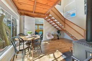 A wooden ceiling, a dining area with chairs, a staircase, and a wood-burning stove.