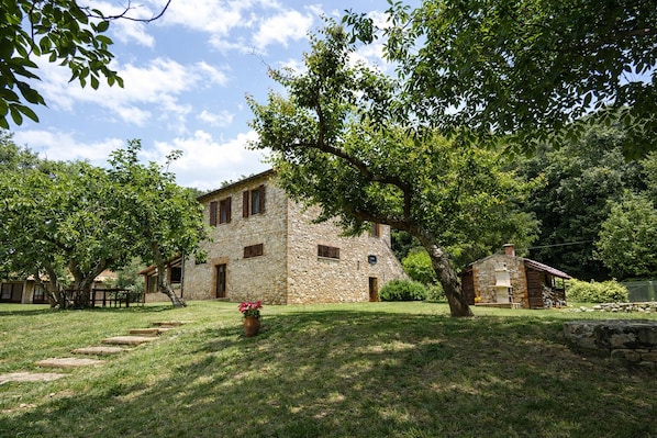 A stone building with a large tree in front, a small wooden structure to the right, and a grassy area with steps leading to the building.