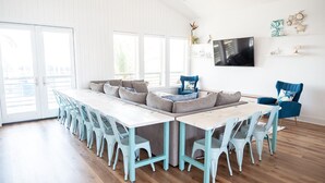 Ein heller, geräumiger Raum mit einem langen Esstisch, blauen Stühlen, einer Couch und einem an der Wand befestigten Fernseher.