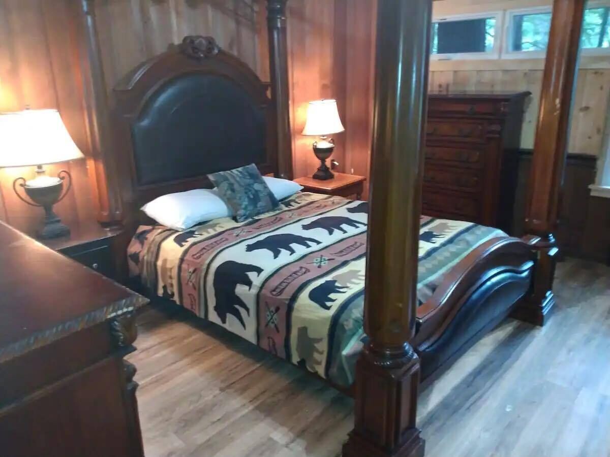 A four-poster bed with a patterned bedspread, two bedside lamps, and a wooden dresser in a room with wooden paneling.