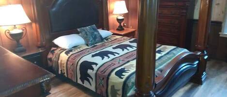 A four-poster bed with a patterned bedspread, two bedside lamps, and a wooden dresser in a room with wooden paneling.