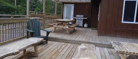 Une terrasse en bois avec des tables et des chaises pour pique-nique, un barbecue et un petit bâtiment doté d’une porte et d’une fenêtre.
