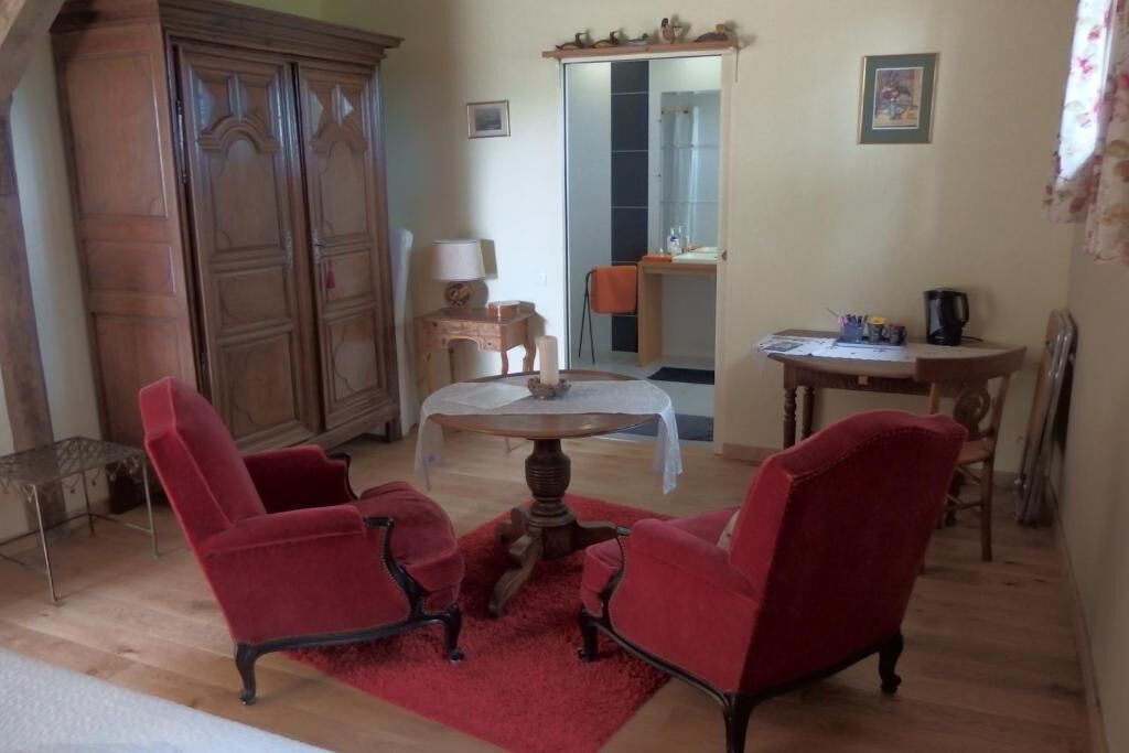Ein Zimmer mit einem Holfischschrank, einem runden Tisch mit rotem Teppich, zwei roten Sesseln, einem kleinen Holznachttisch und einem Spiegel über einer Tür.