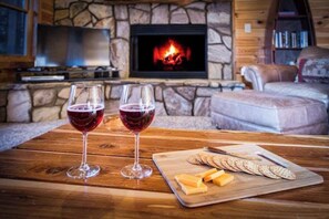 Un salon chaleureux avec une cheminée en pierre, un téléviseur et une table en bois sur laquelle sont disposés des verres de vin rouge et une planche de fromages.