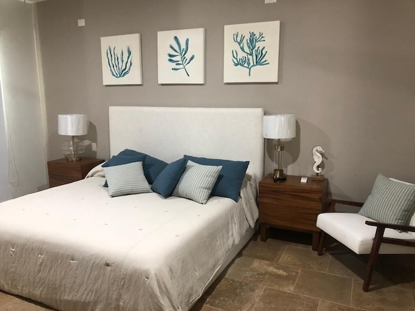 Un dormitorio con una cama grande, dos mesitas de noche con lámparas, una silla y tres obras de arte colgadas en la pared.