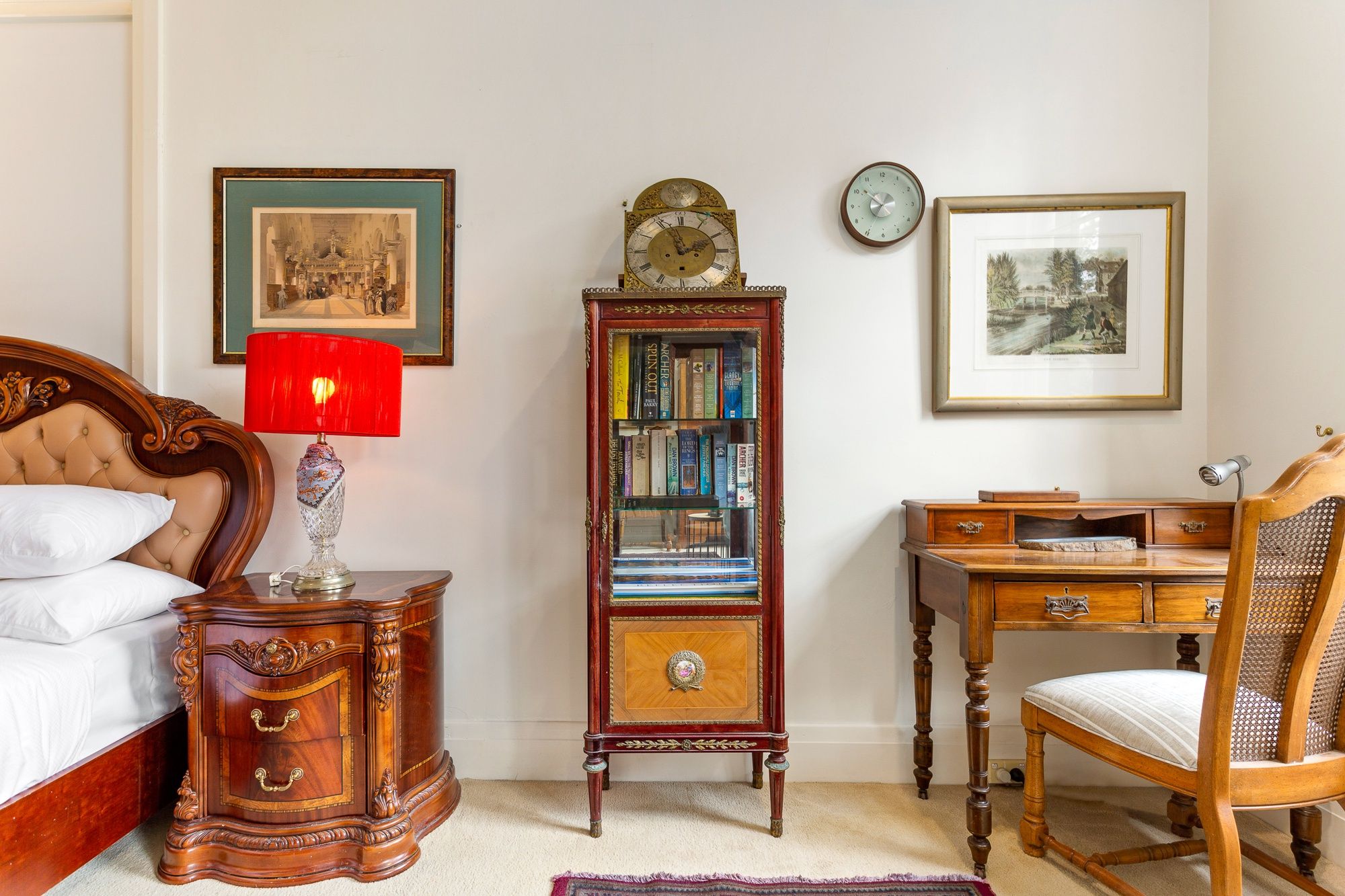 A bedroom with a bed, a nightstand with a lamp, a bookshelf, a desk, a chair, and two framed pictures on the wall.