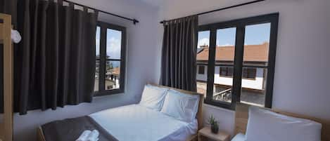 A room with two beds, large windows, a hanging light fixture, and a view of buildings outside.