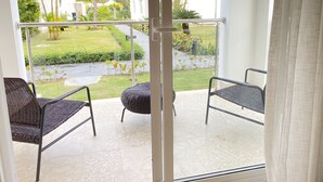 A balcony with two wicker chairs and a small round ottoman, overlooking a well-maintained garden with palm trees and a paved path.