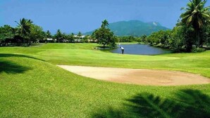 A golf course with a pond, palm trees, and mountains in the background.