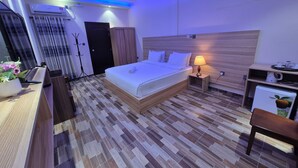 Una habitación de hotel moderna con una cama bien hecha, una lámpara de noche, un pequeño escritorio con una silla y un refrigerador.