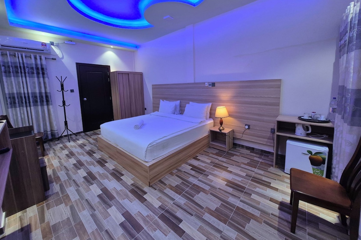 A hotel room with a bed, bedside table, lamp, and a small kitchenette.