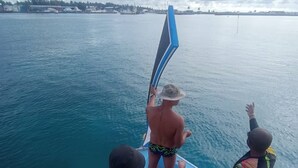Un bote con cubierta de madera y barandilla azul, con tres personas de pie al borde, una sosteniendo una tabla de surf.