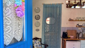 A cozy room with a blue door, a wooden chair, a small table, a refrigerator, and a kitchen area with a window and curtains. - Casal DOS Carpinteiros 4 (Silveira)