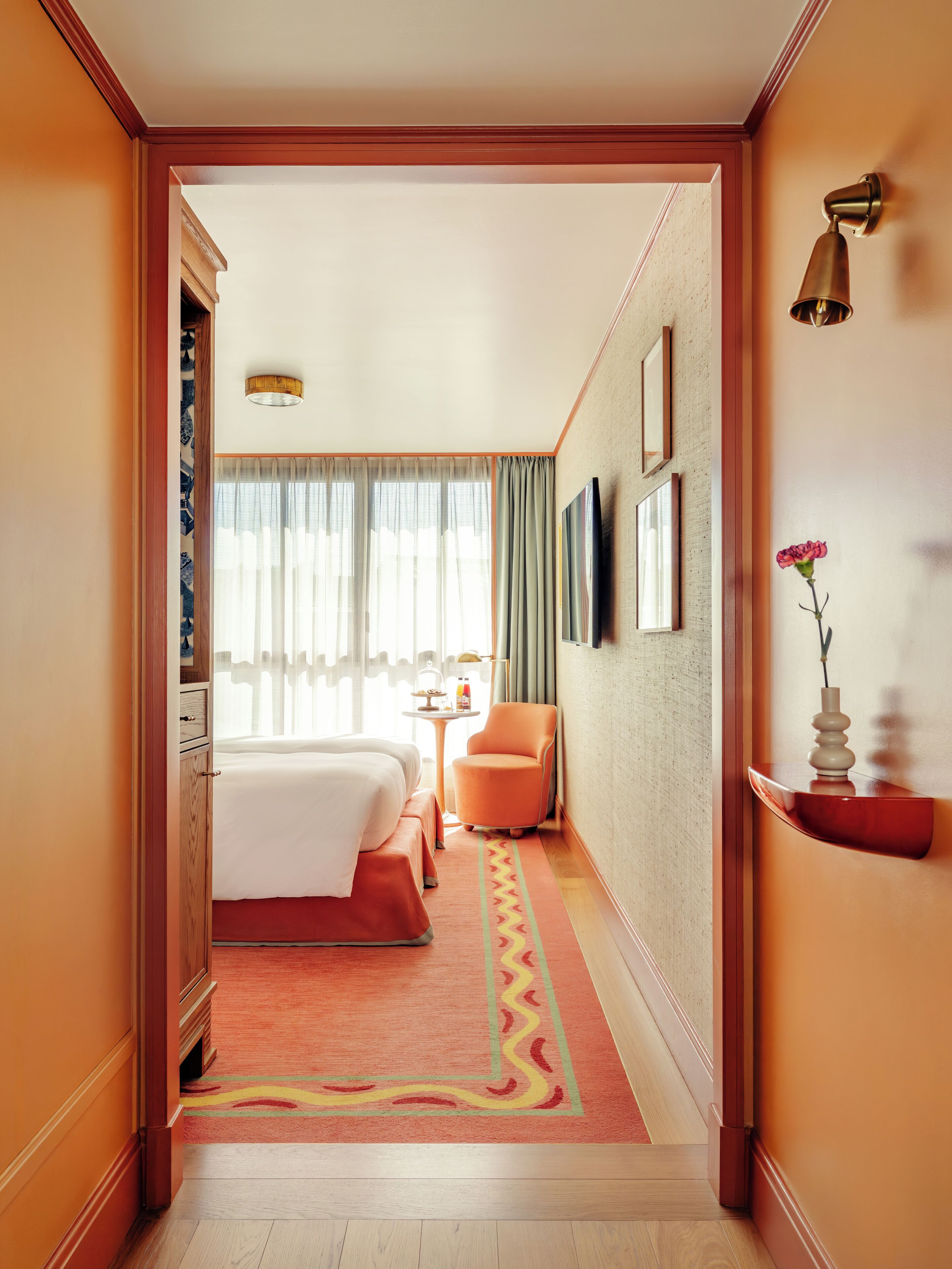 Una camera d'hotel con un letto, una sedia, un tavolino, un vaso con un fiore e un televisore a parete.