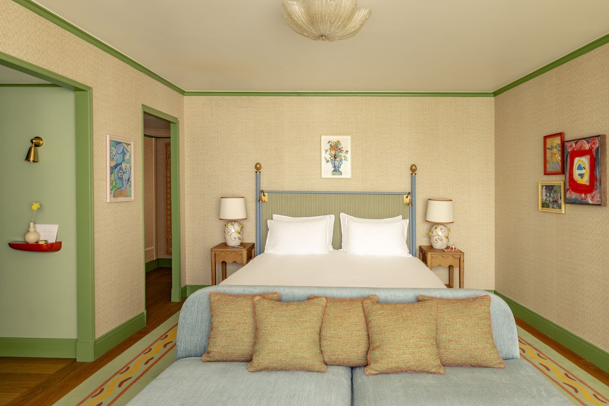 A hotel room with a large bed, two bedside tables with lamps, a sofa with cushions, a door with a green frame, and a wall with framed artwork.