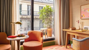 A cozy hotel room with a large window, a potted plant, two chairs, a small table with a lamp, and a desk with a chair. - La Fantaisie (Paris)
