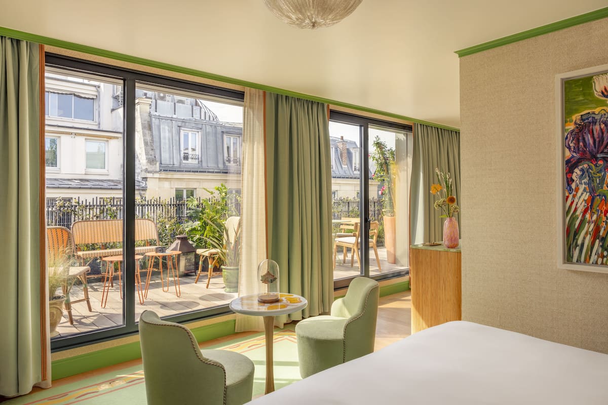 A hotel room with a large window, a bed, two chairs, a small table with a vase, and a painting on the wall.