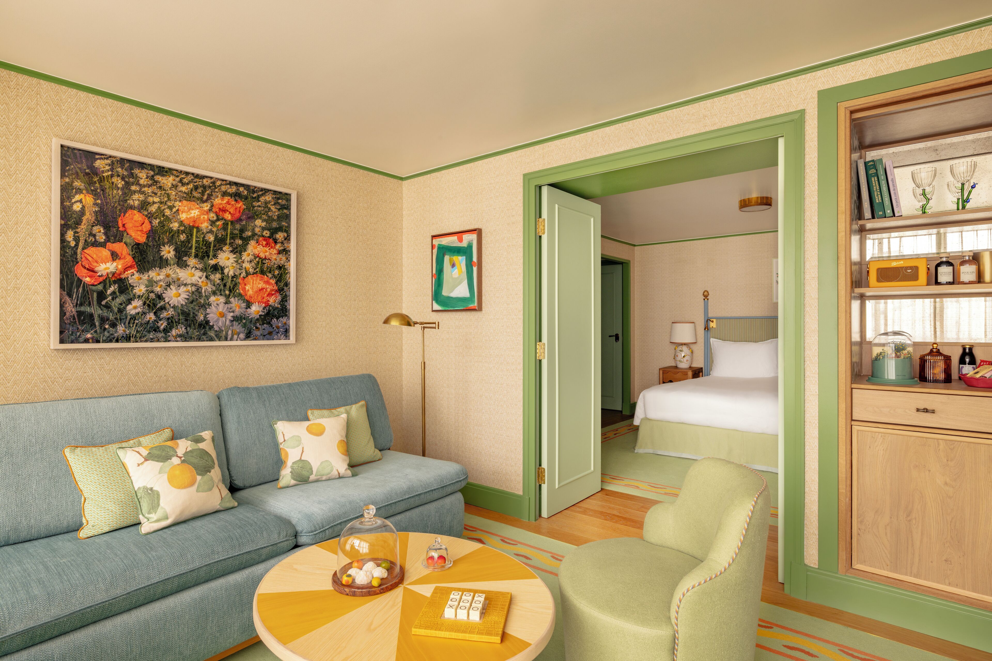 A hotel room with a blue sofa, a green armchair, a wooden cabinet with decorative items, and a painting of flowers on the wall.