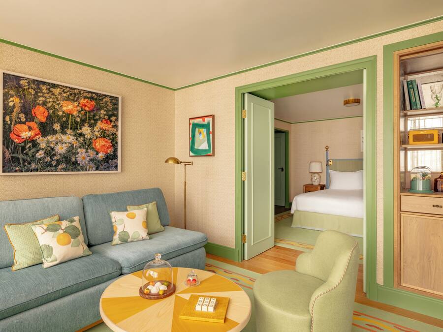 A hotel room with a blue sofa, a green armchair, a wooden cabinet with decorative items, and a painting of flowers on the wall.