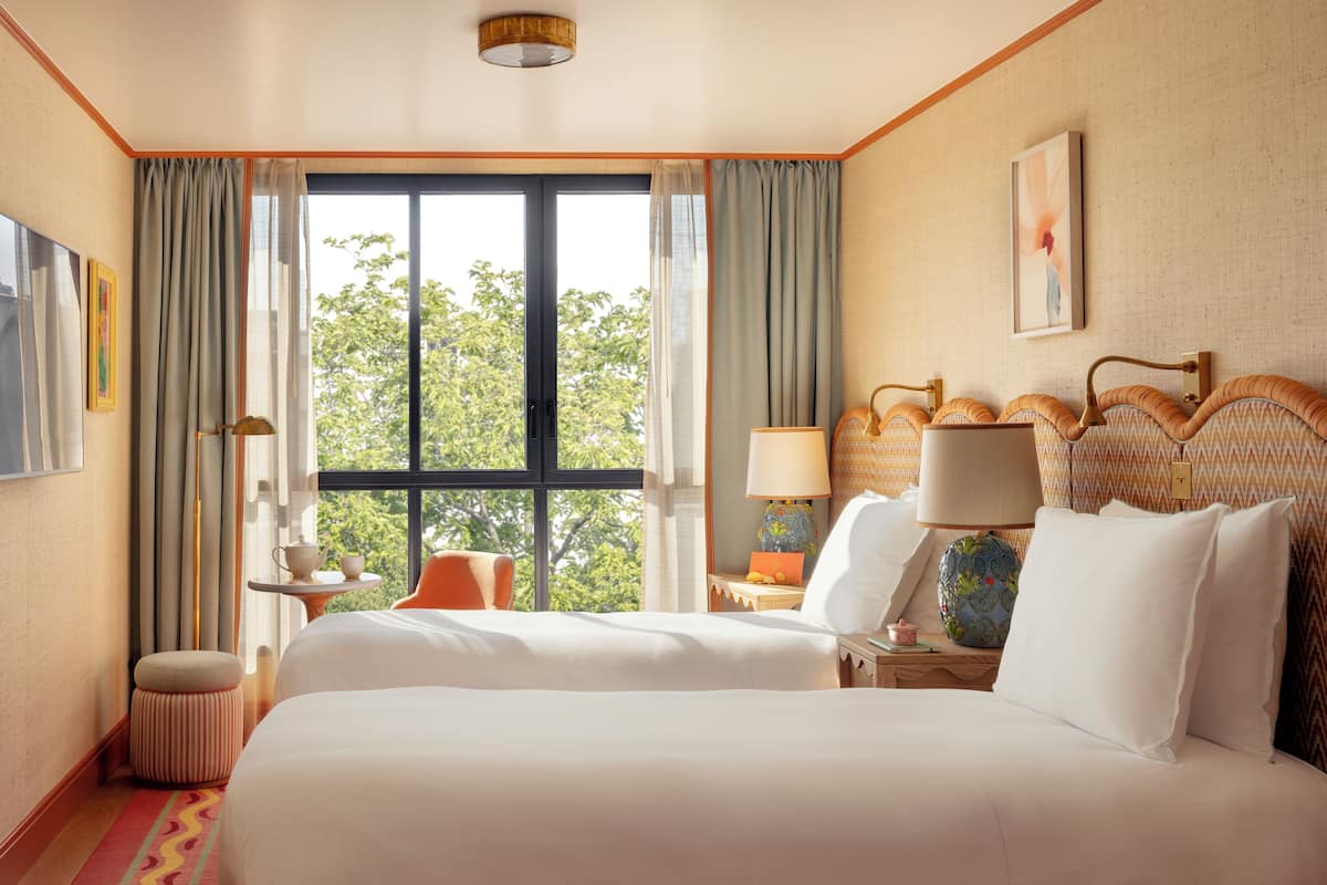 A hotel room with a large bed, a nightstand with a lamp, a small table with a cup, and a view of trees through the window.