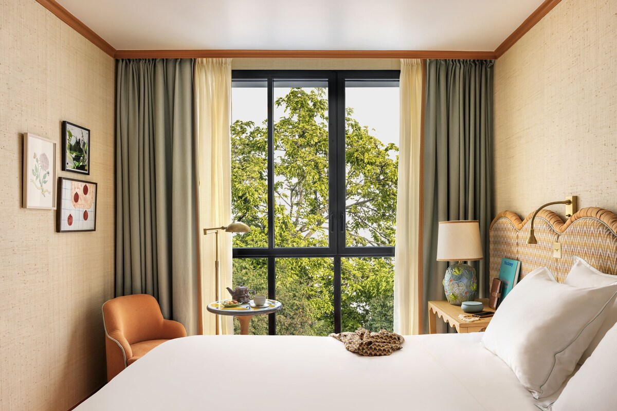 A hotel room with a large bed, a chair, a small table with a lamp, and a view of trees through the window.