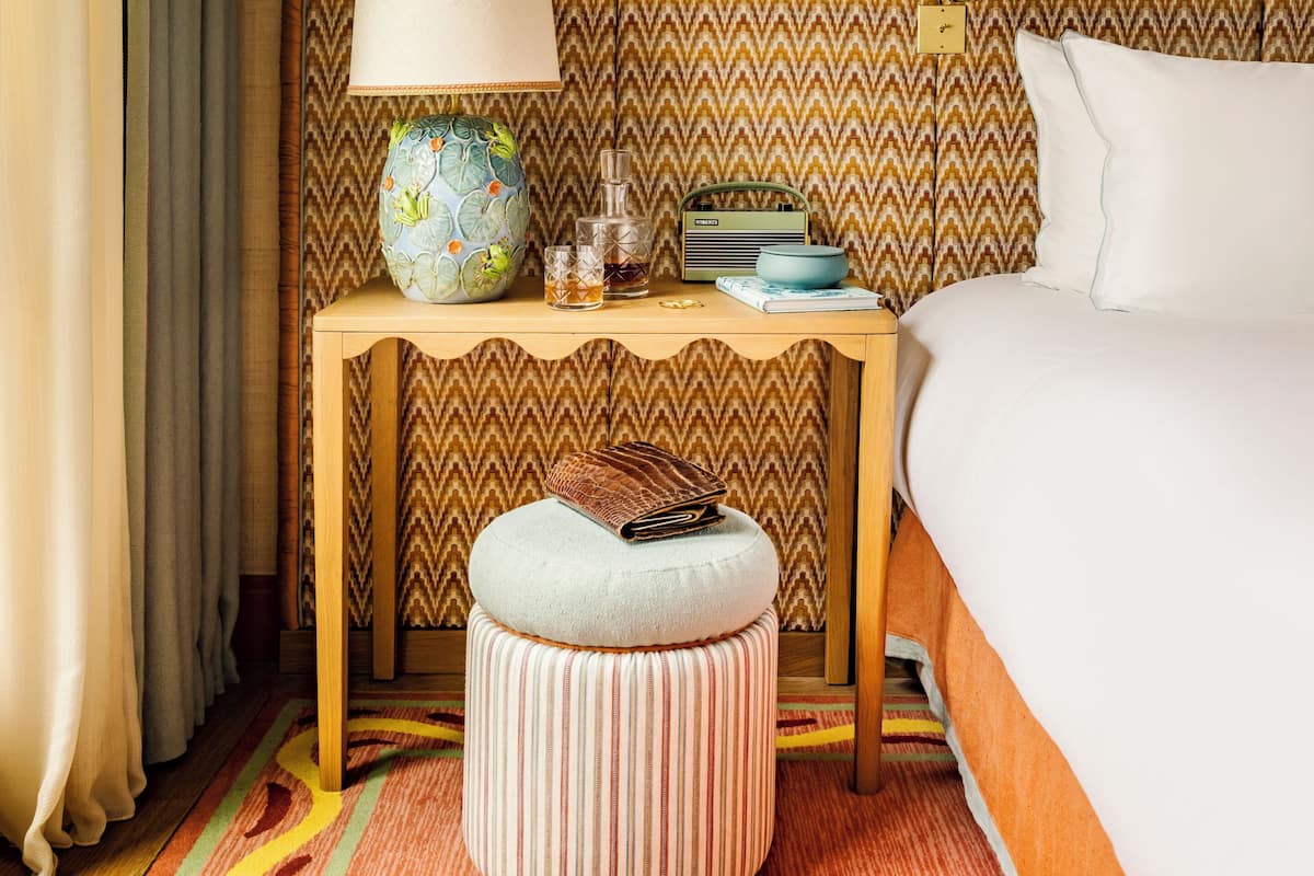 A hotel room with a bed, a nightstand, a lamp, a radio, a vase, and a striped ottoman.