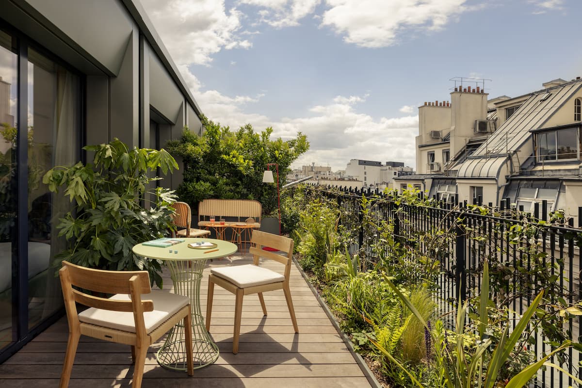 A rooftop terrace with a wooden table and chairs, surrounded by lush greenery and a black metal fence.