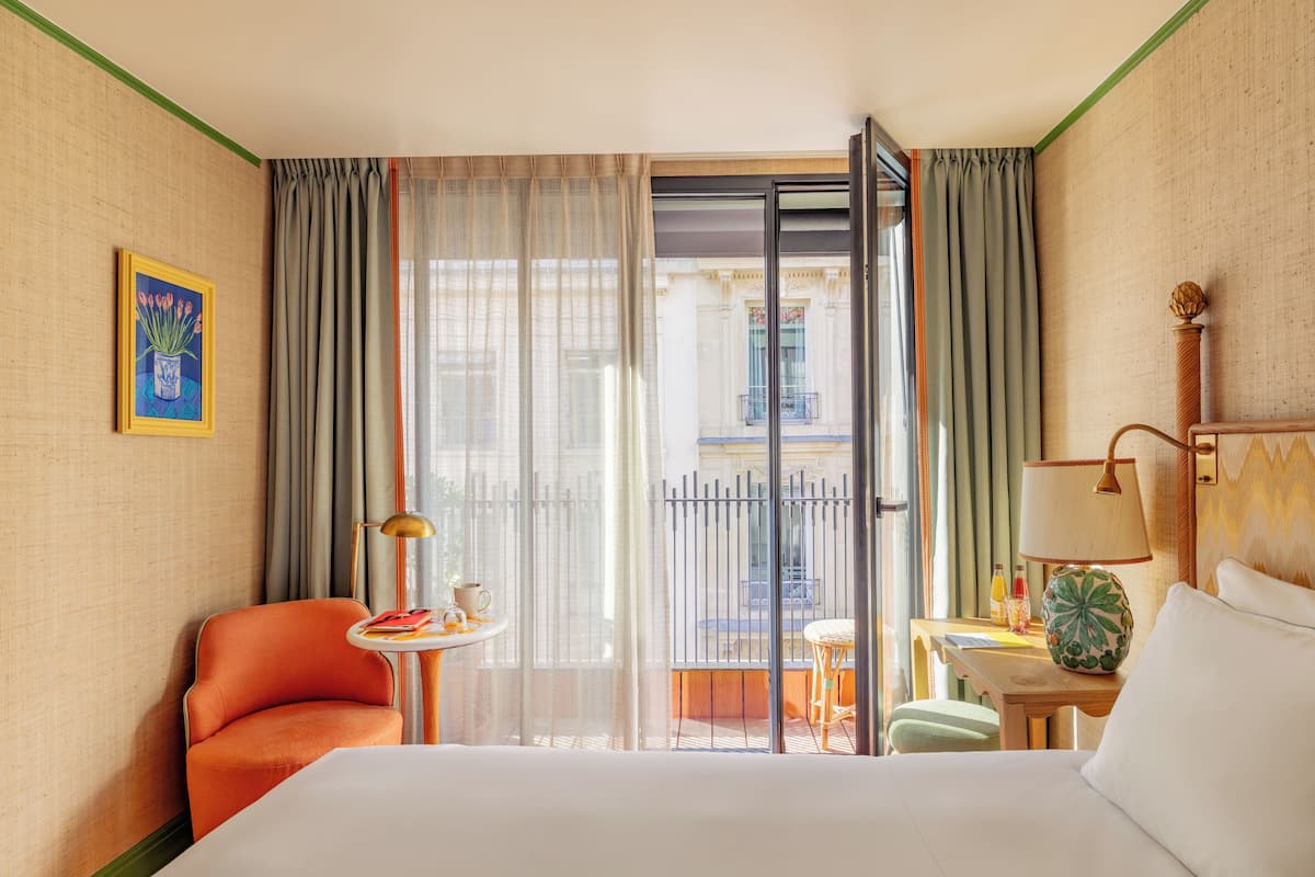 A hotel room with a bed, a chair, a small table with a lamp, and a view of the outside through a large window.