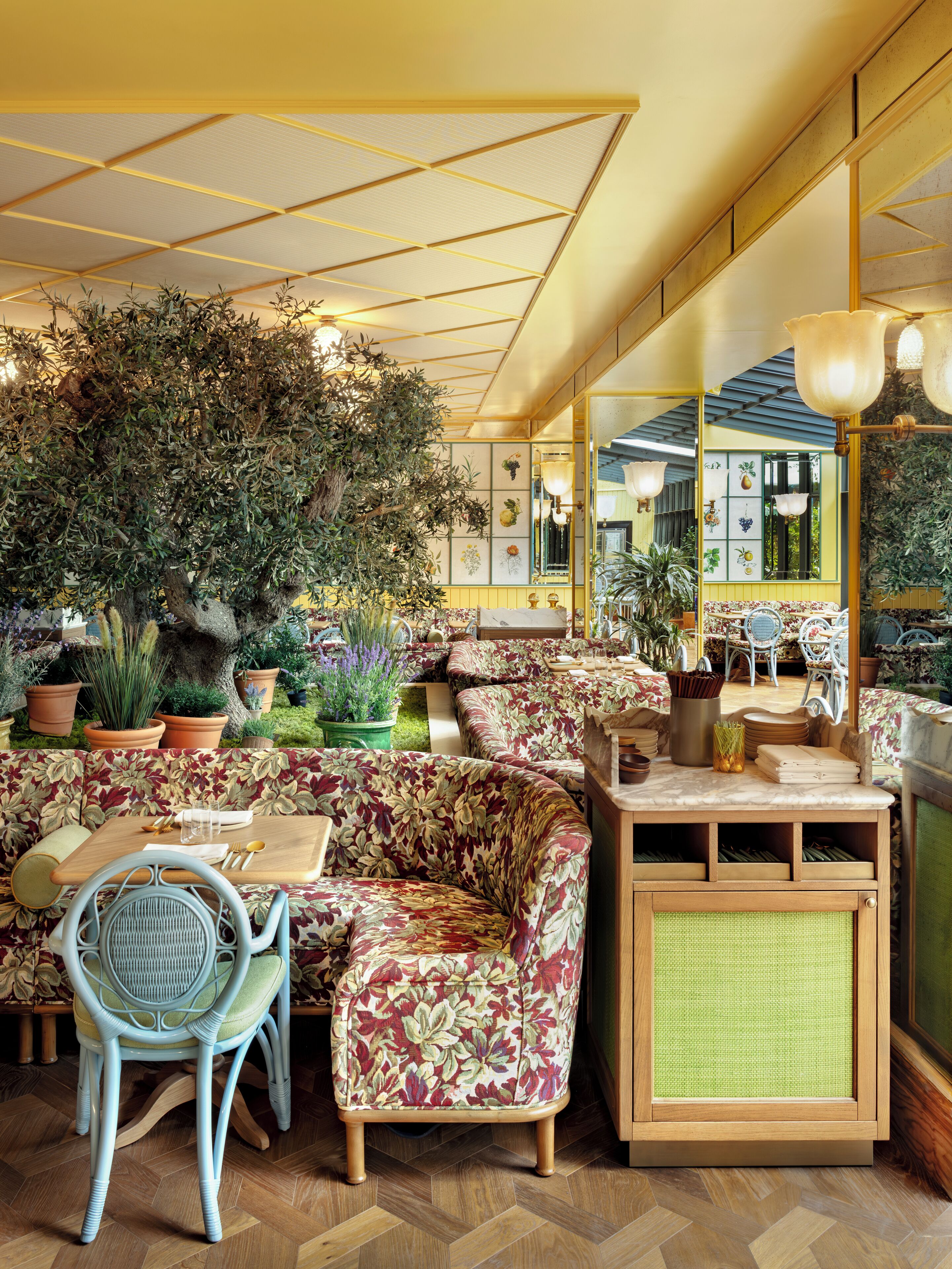 A cozy indoor seating area with a sofa, chairs, and a table, surrounded by potted plants and a large mirror reflecting the room.