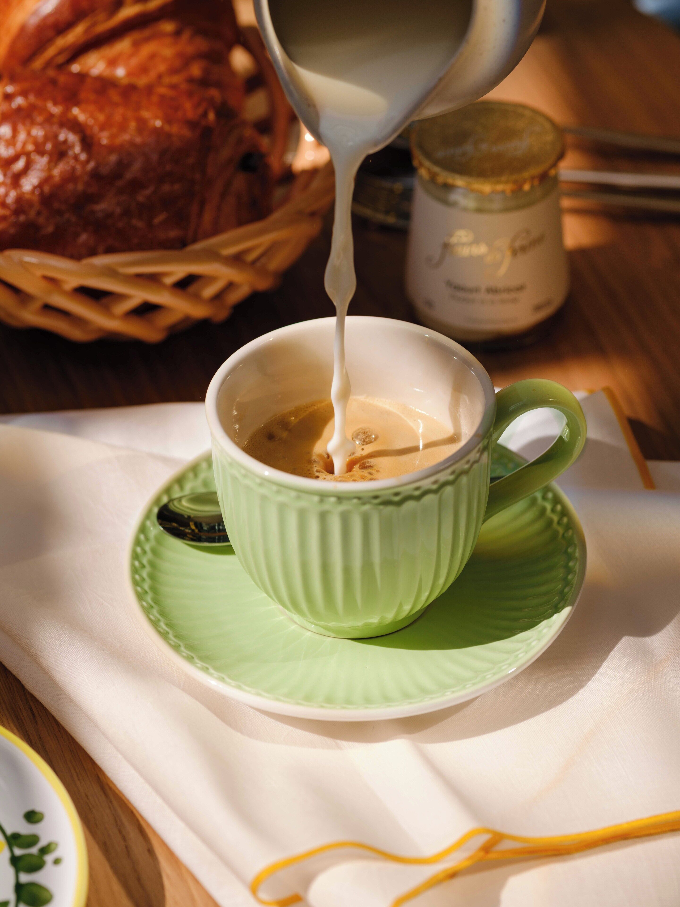 A cup of coffee with milk being poured into it, placed on a green saucer.