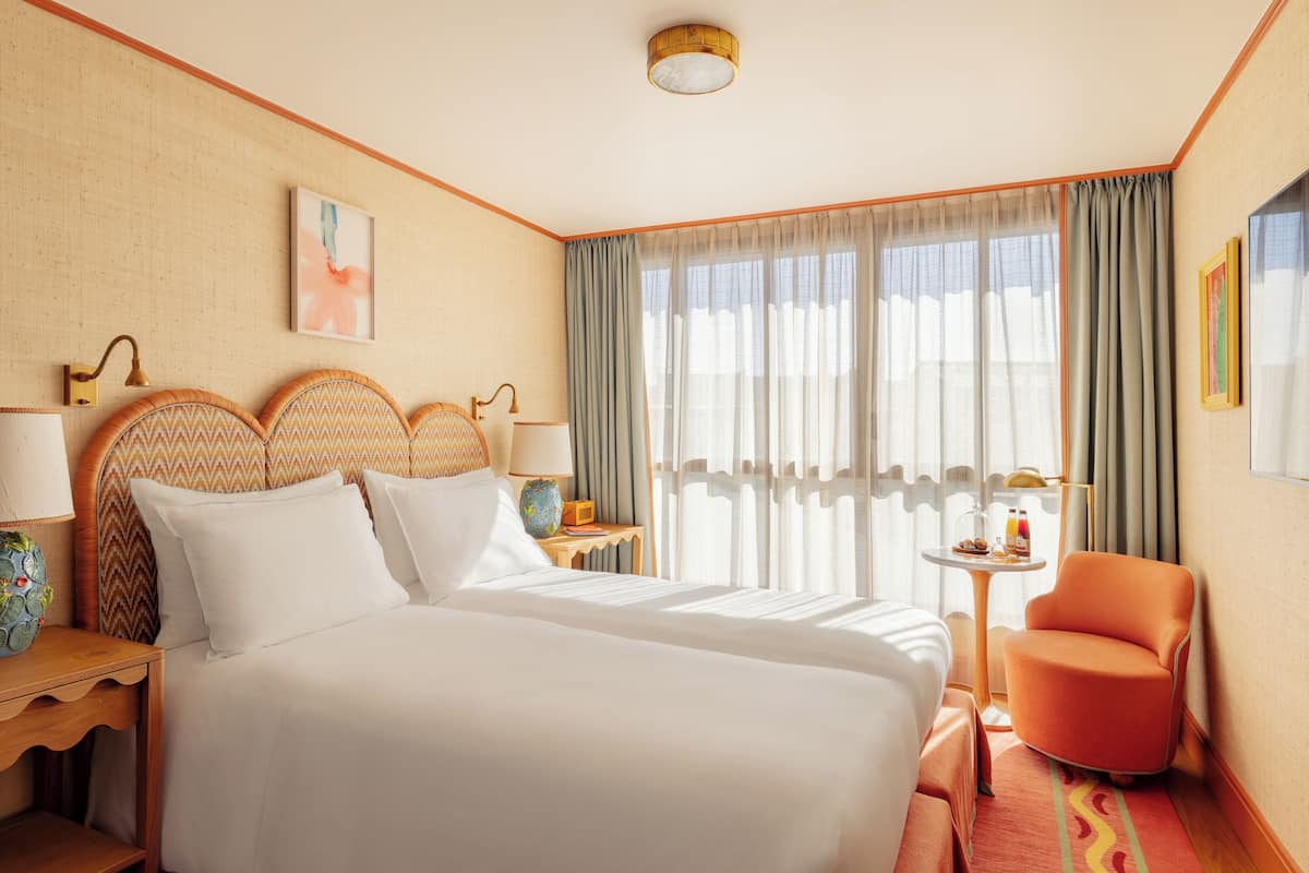 A hotel room with a large bed, two bedside tables, a chair, a small table with a lamp, and a window with curtains.