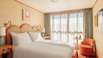 Habitación de hotel con una cama grande, dos mesitas de noche, una silla, una pequeña mesa con lámpara y una ventana con cortinas.
