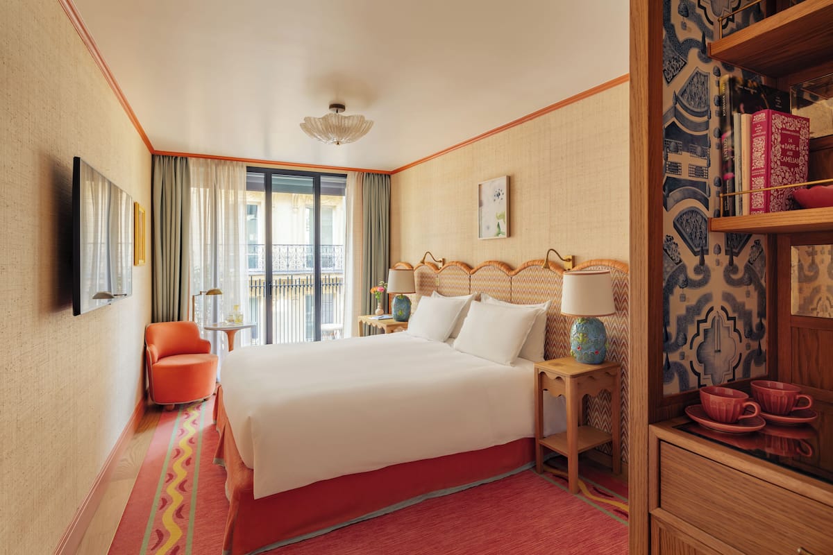A hotel room with a large bed, a sitting area with a chair, a small table with a lamp, and a bookshelf with books and decorative items.