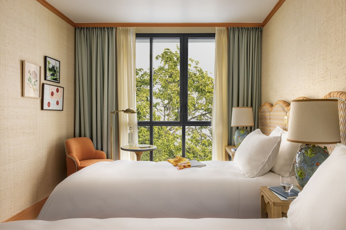 A hotel room with a large bed, a chair, a small table, a lamp, and a view of trees through the window.