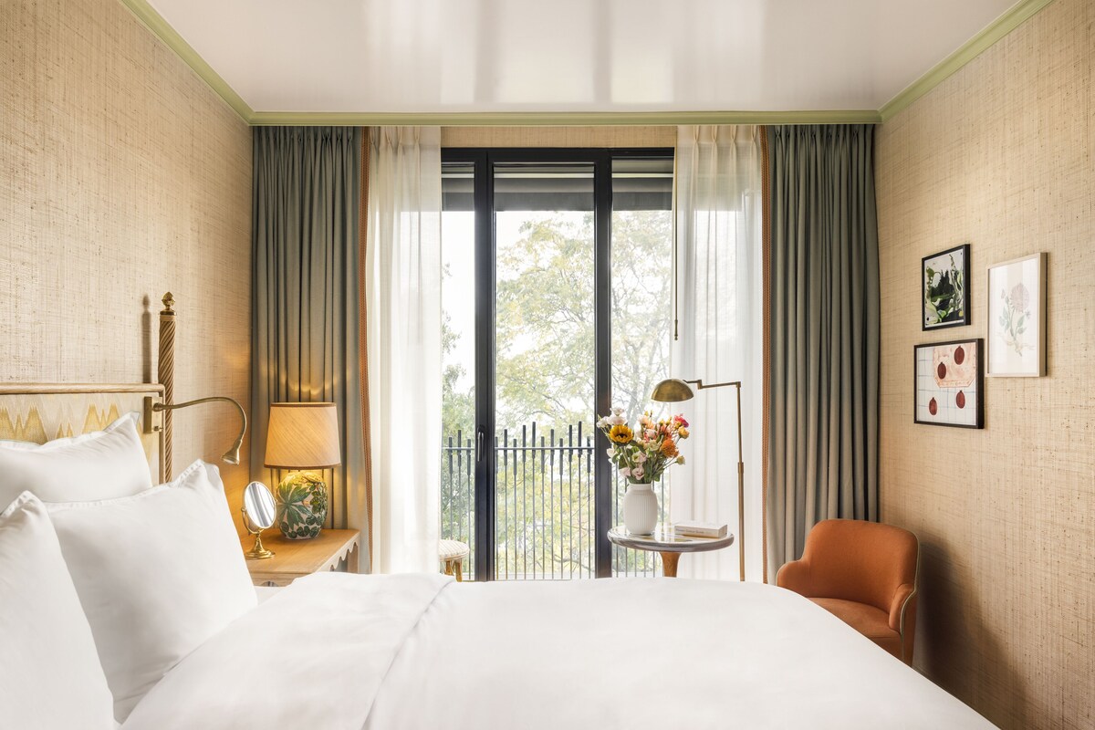 A hotel room with a bed, a chair, a nightstand, a lamp, a vase with flowers, and a window with curtains.