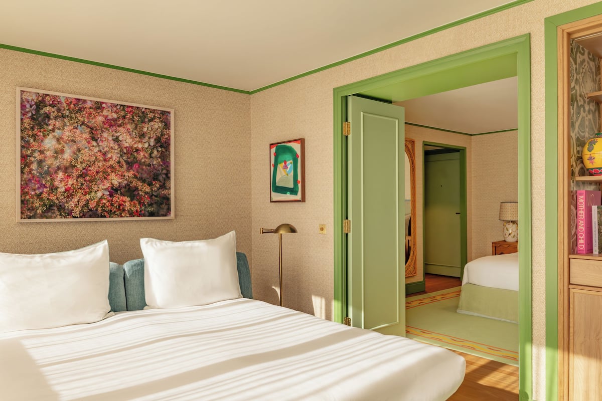 A hotel room with a large bed, a painting on the wall, and a green-framed door leading to another room.