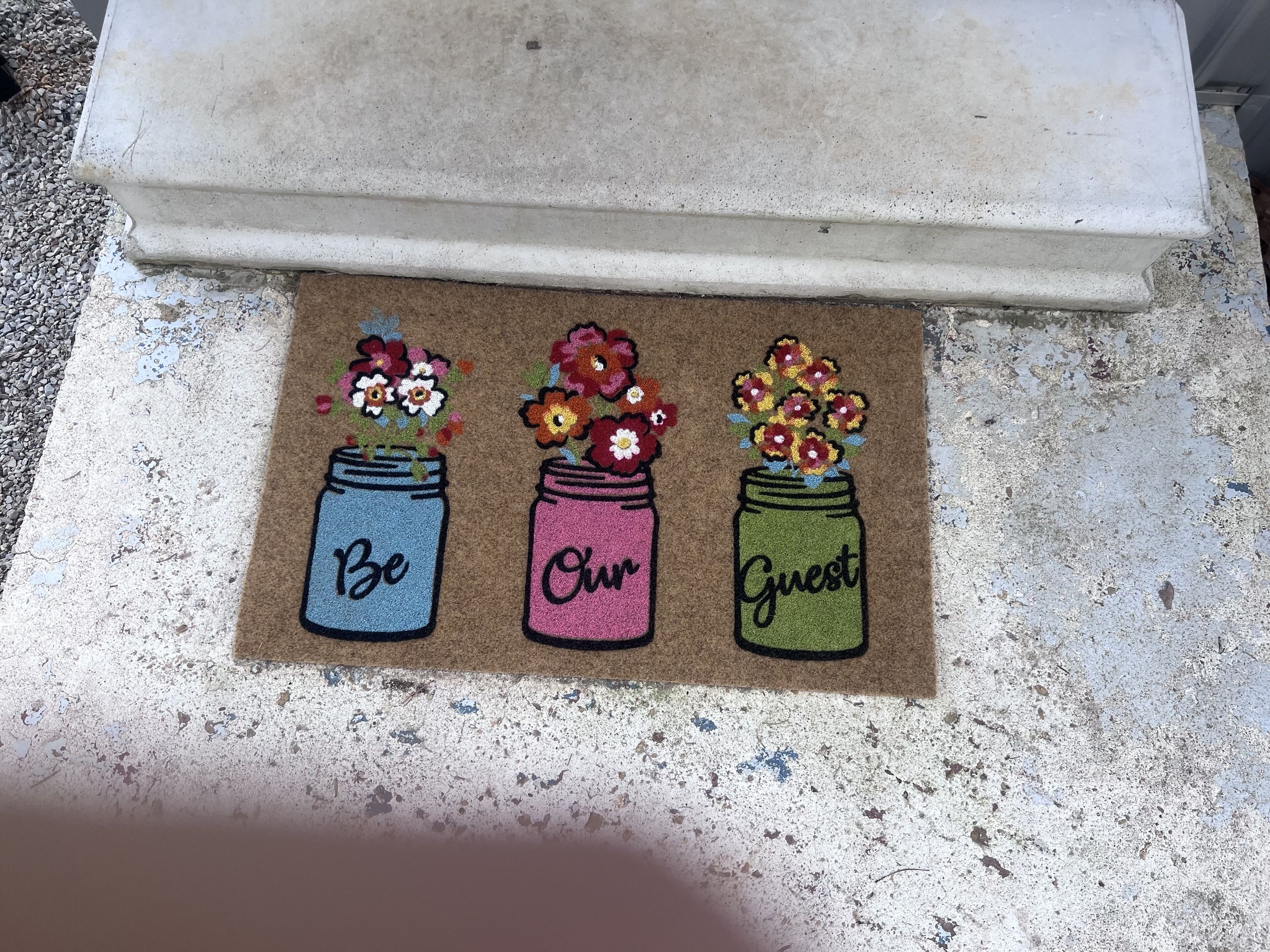 A doormat with three jars of flowers and the words "Be Our Guest" on it.