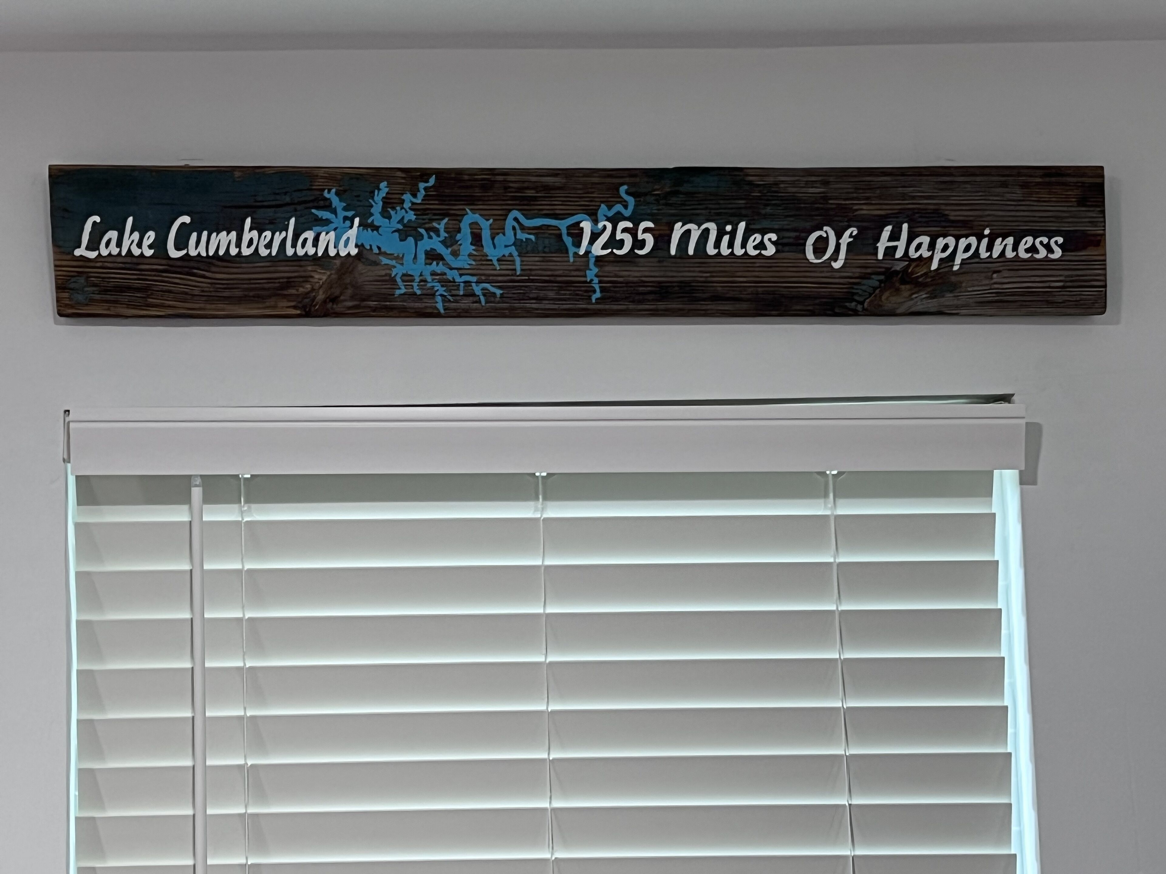 Une enseigne en bois, avec du texte et un motif abstrait bleu, fixée au-dessus d’une fenêtre munie de stores horizontaux.