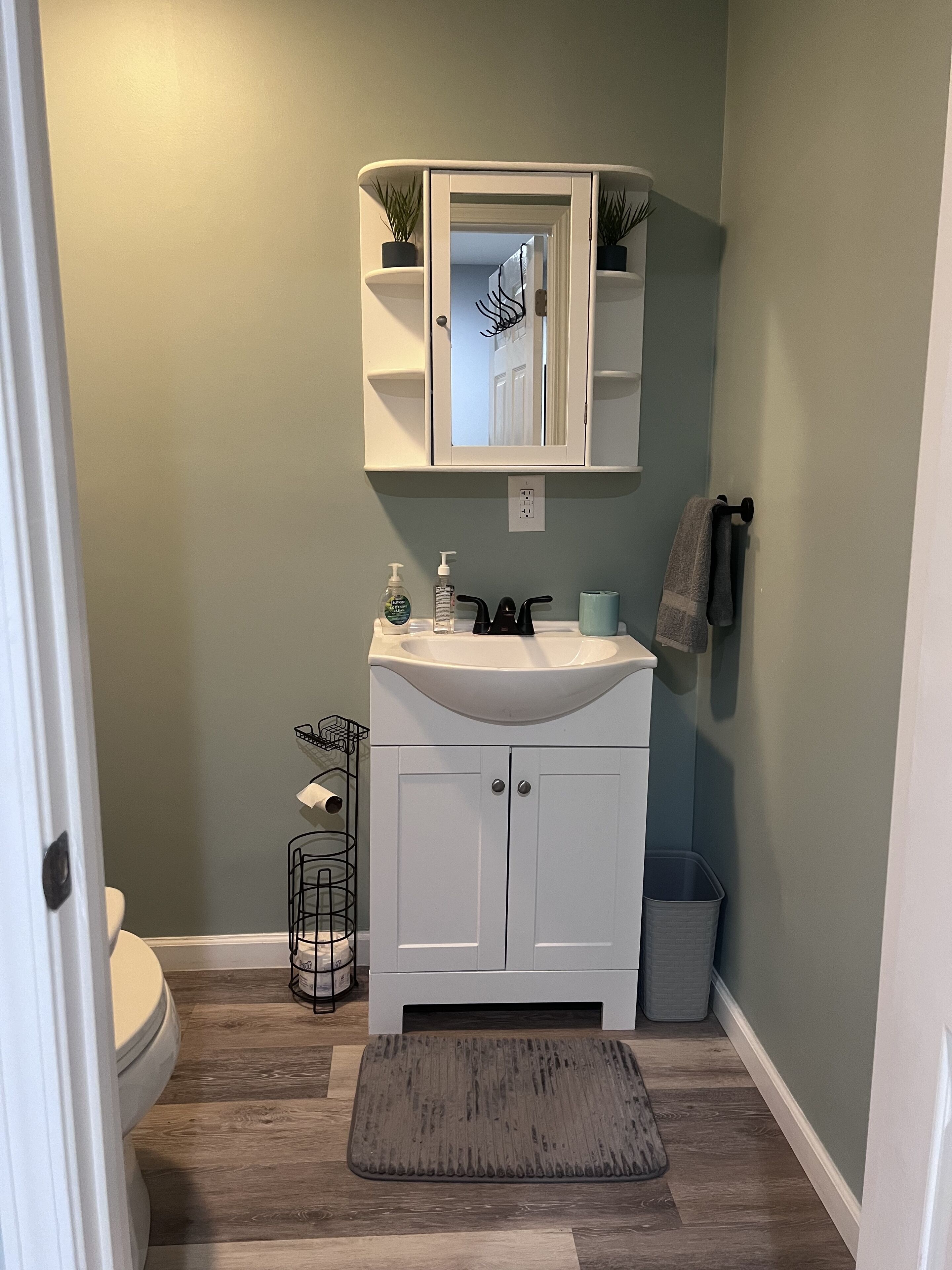 Une salle de bain avec un lavabo blanc, un miroir au-dessus, et une serviette accrochée au mur. On y trouve également une poubelle et une petite plante posée sur le sol.