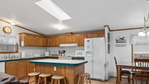 Une cuisine avec des meubles de rangement en bois, un îlot central doté de tabourets, une table à manger avec des chaises et un réfrigérateur.