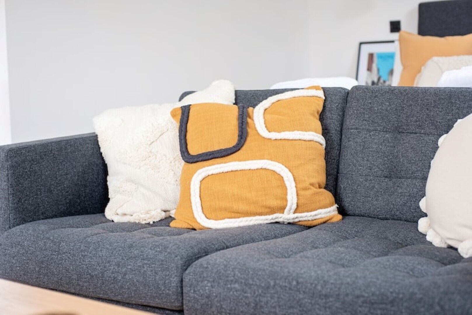 A grey sectional sofa with three cushions: a white one, a mustard-colored one with a black outline, and a white one with a fluffy texture.