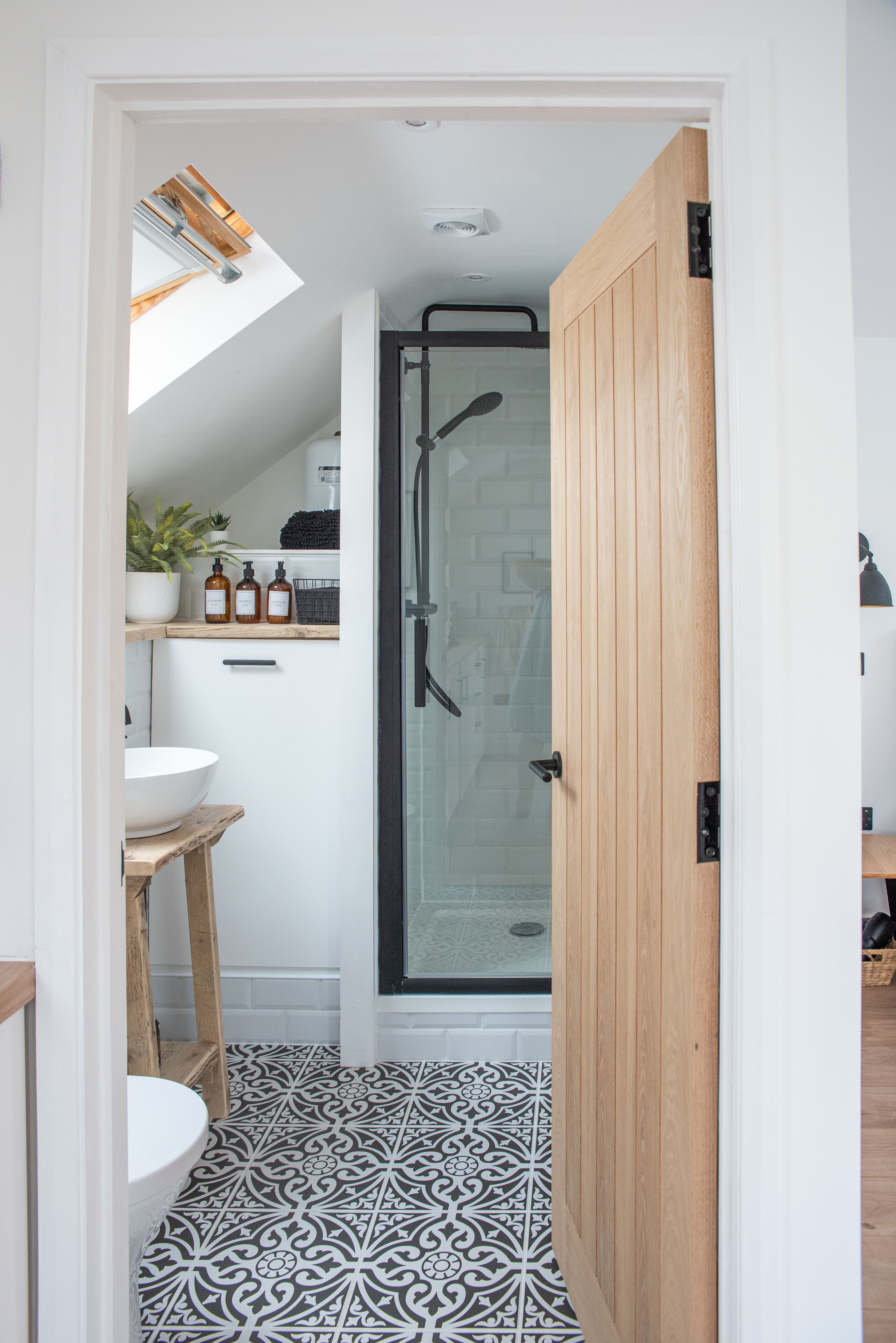 A bathroom with a white sink, wooden stool, black and white patterned floor, a glass shower enclosure, and a skylight above.