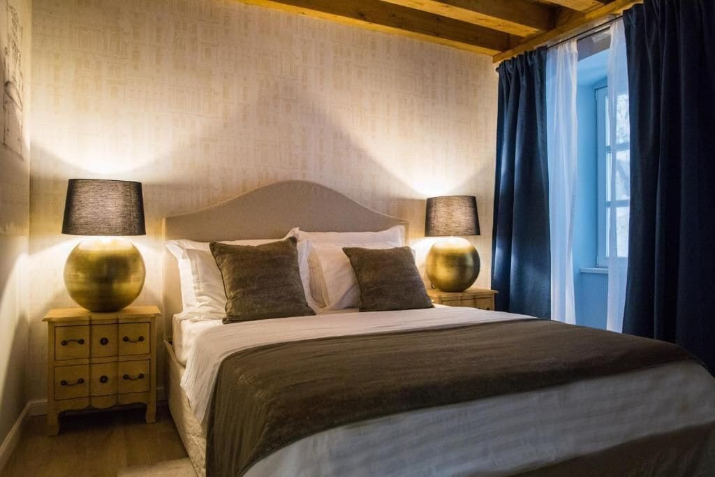 Uma cama bem arrumada com cabeceira, duas mesas de cabeceira com abajures e uma janela com cortinas.